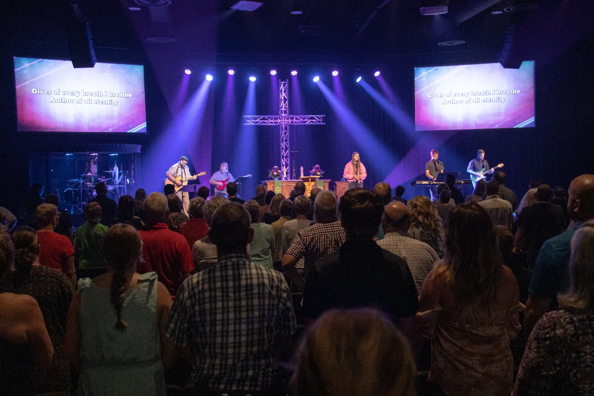 Congregation watching a worship band perform on stage with cross and purple lighting.