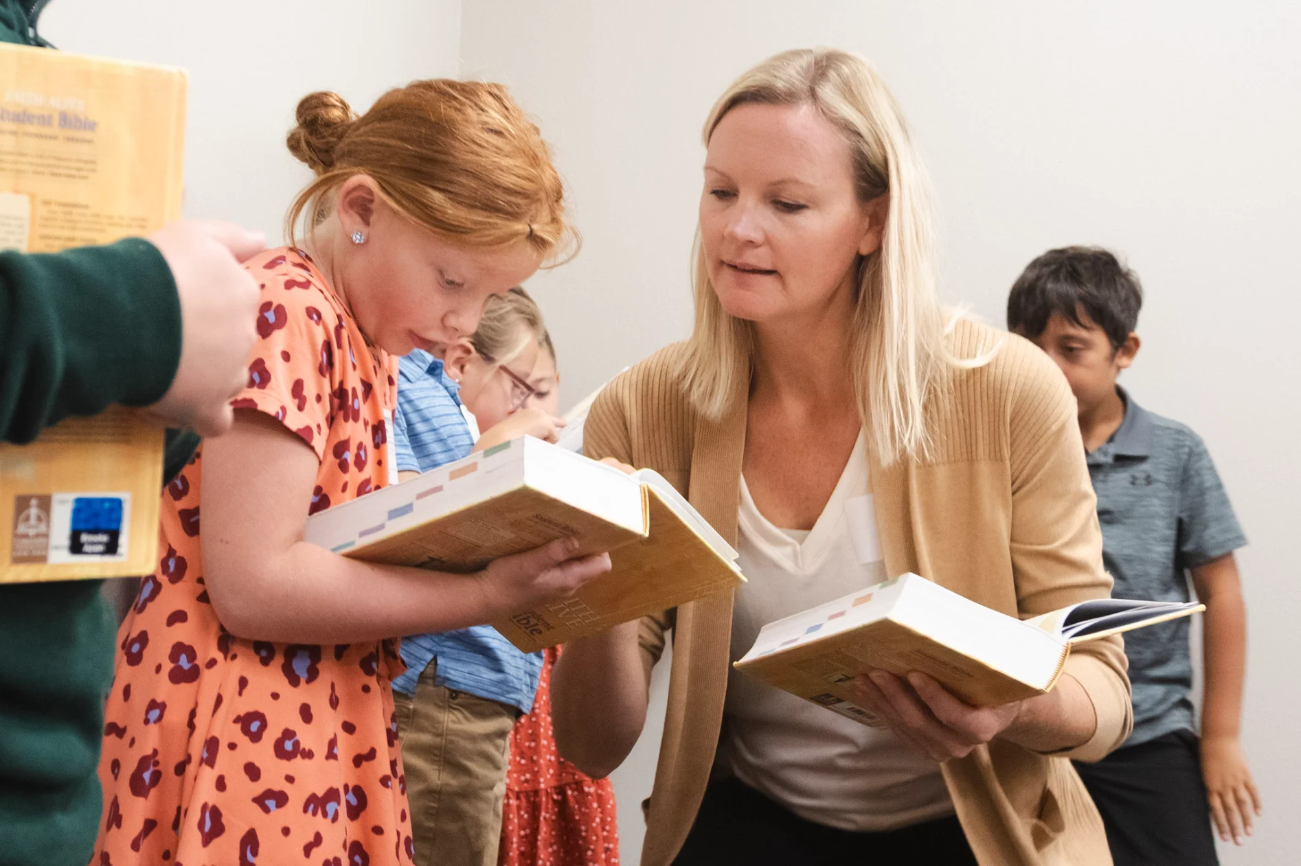 Woman teaching young girl to read from a large book, with children reading in the background.