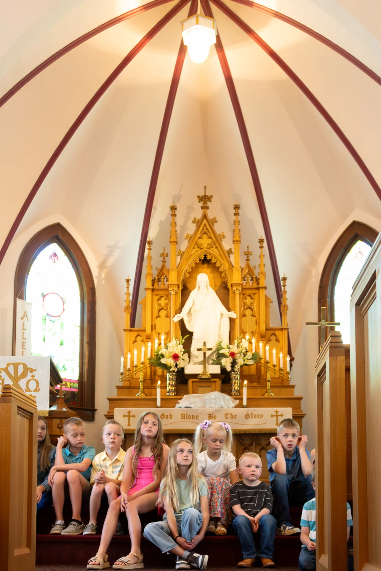 Group of children sitting on steps inside a church with a golden altar and statue behind them.
