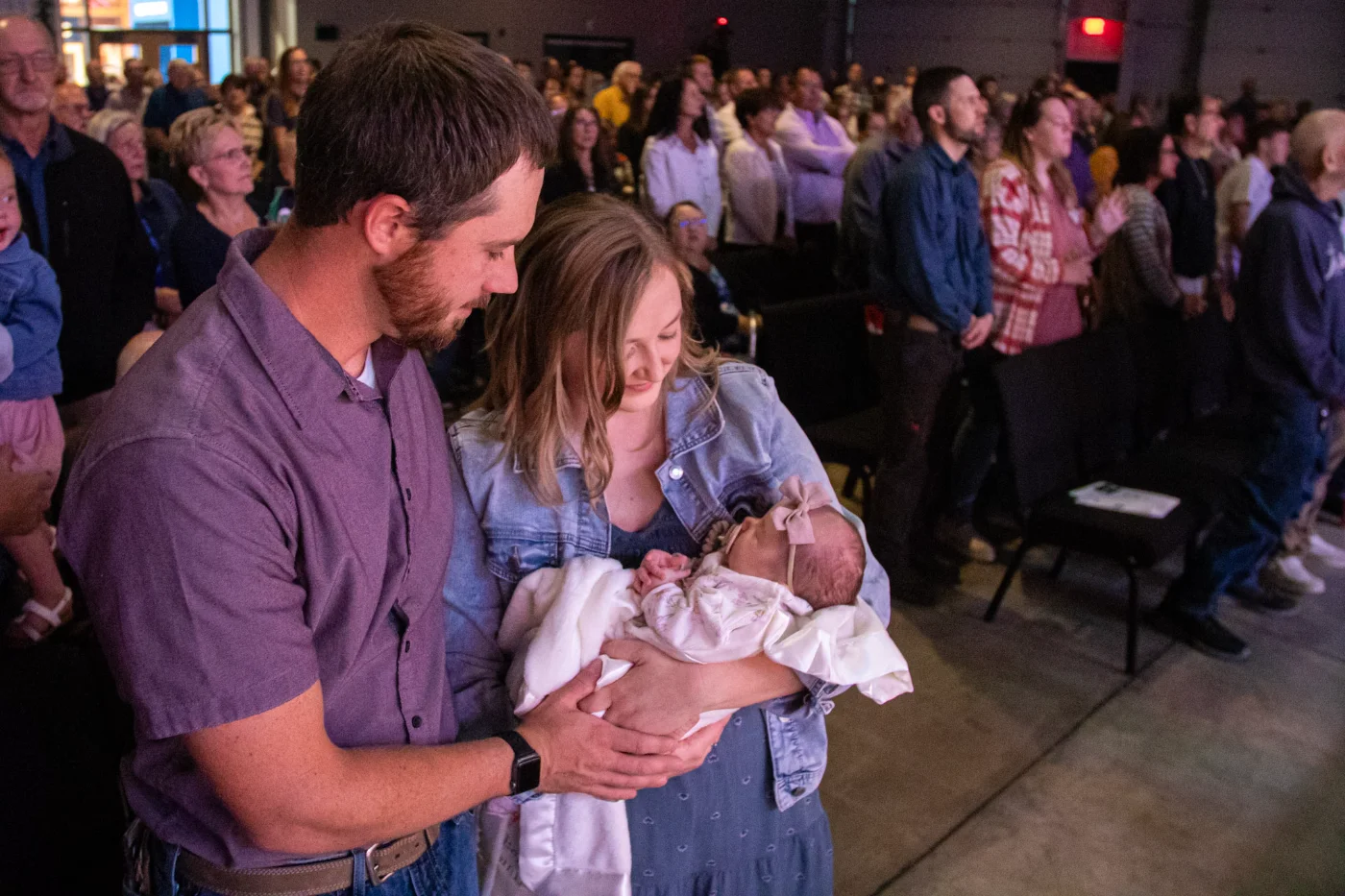 Man and woman holding a newborn baby during a dedication service with seated crowd in background.