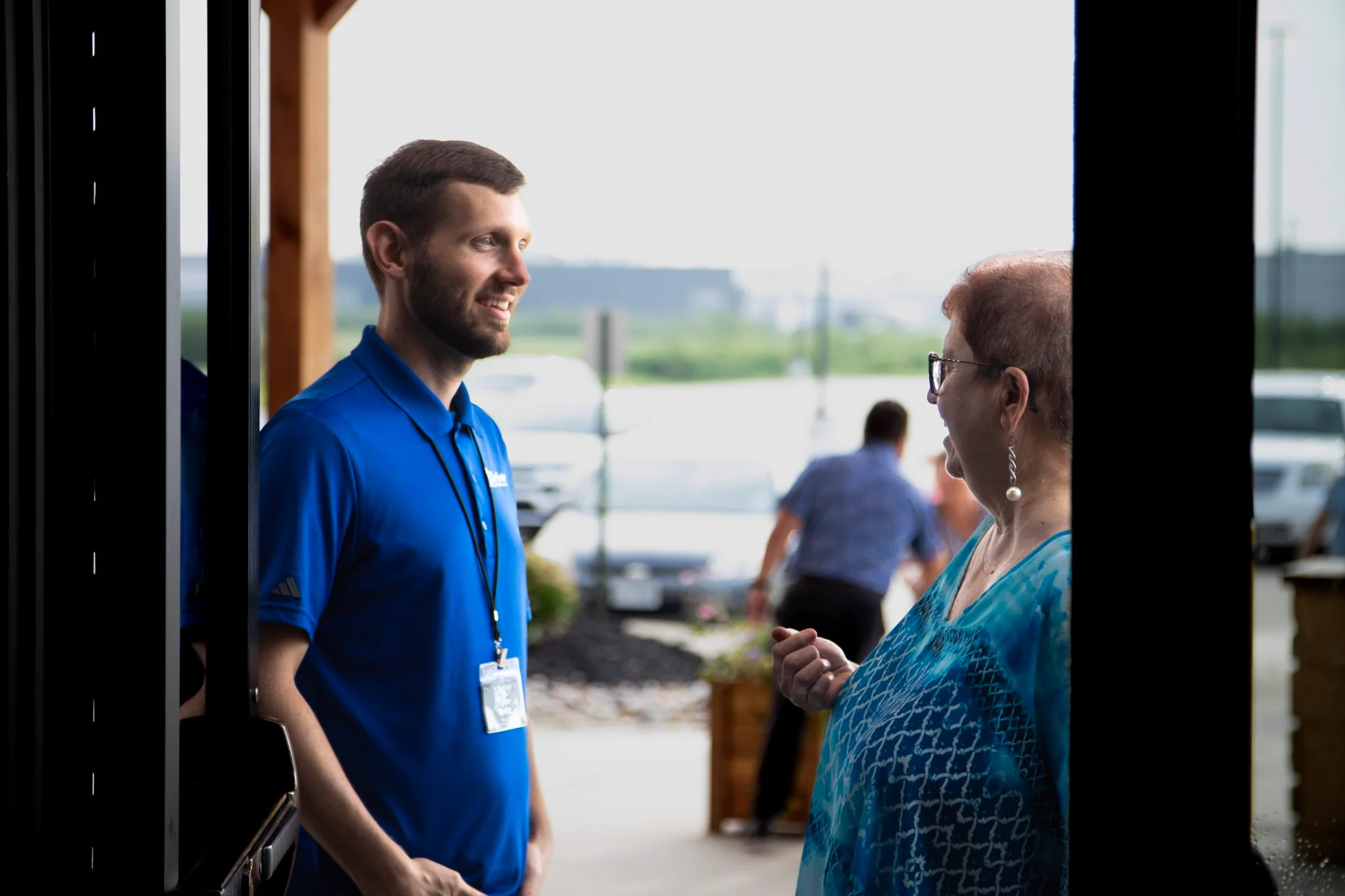 Man in blue shirt warmly greets an older woman wearing glasses outside a building.