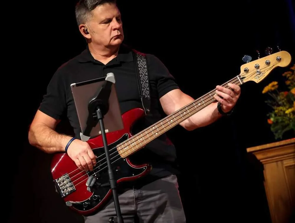 Man playing a red electric bass guitar on stage with a music stand and dark background.