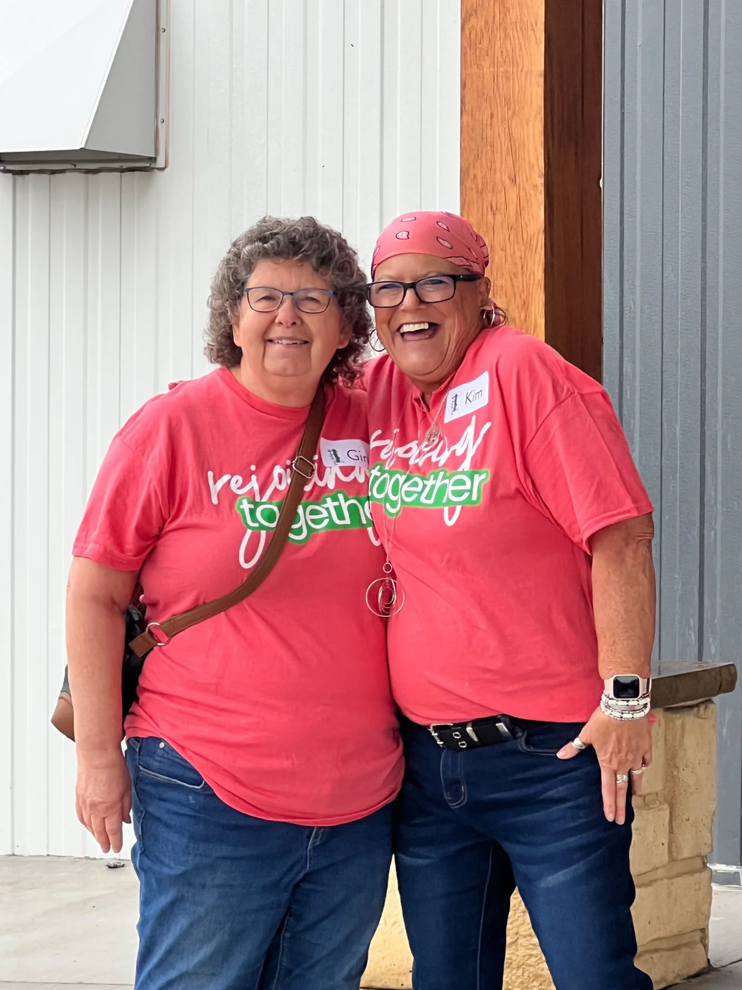 Two smiling women wearing matching red 'rejoicing together' shirts, standing outdoors with arms around each other.