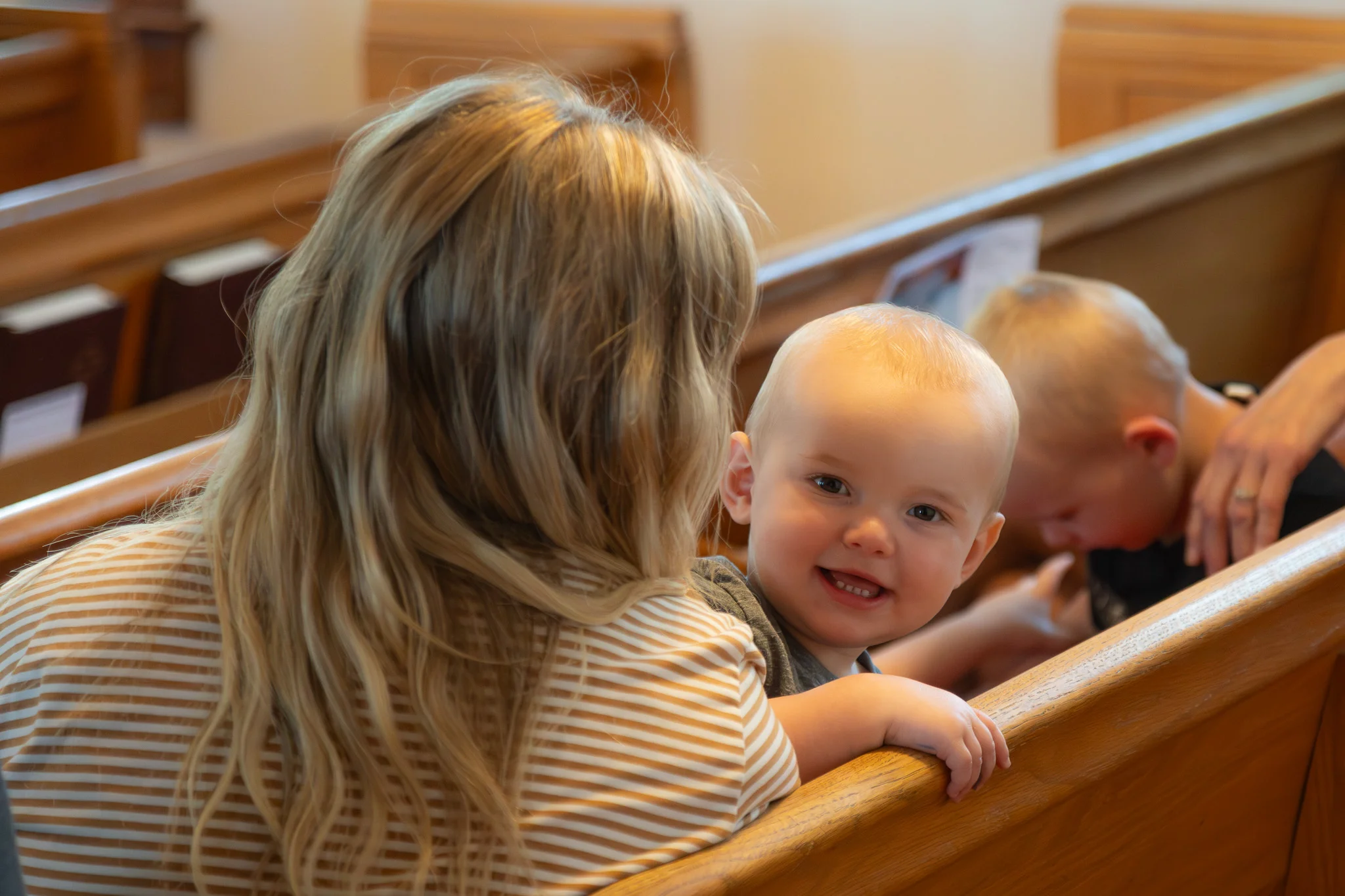 Smiling baby held by a woman sitting in a wooden church pew with another child nearby.