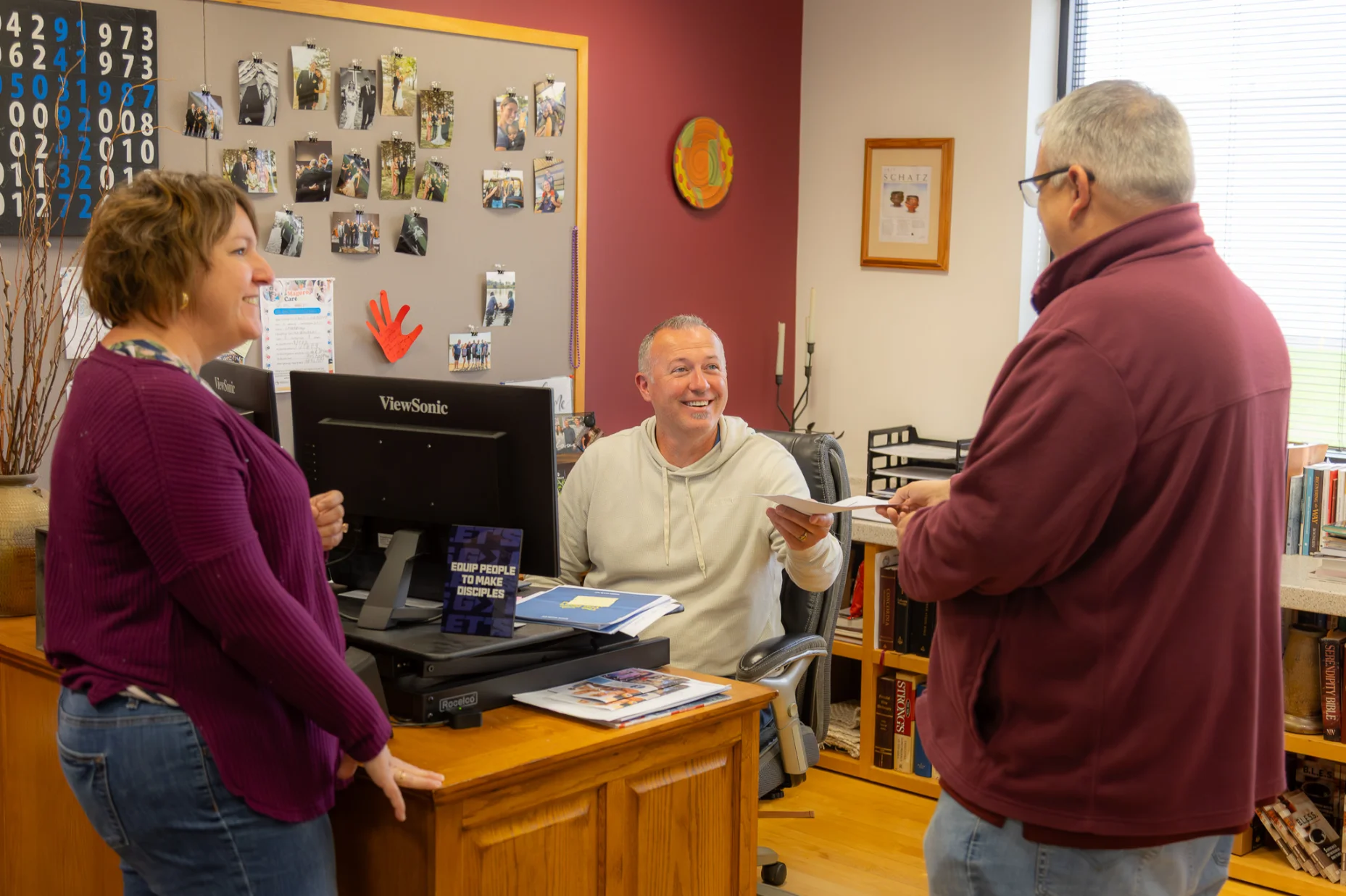 Three people in an office: a man at desk handing paper to another man while a woman watches smiling.