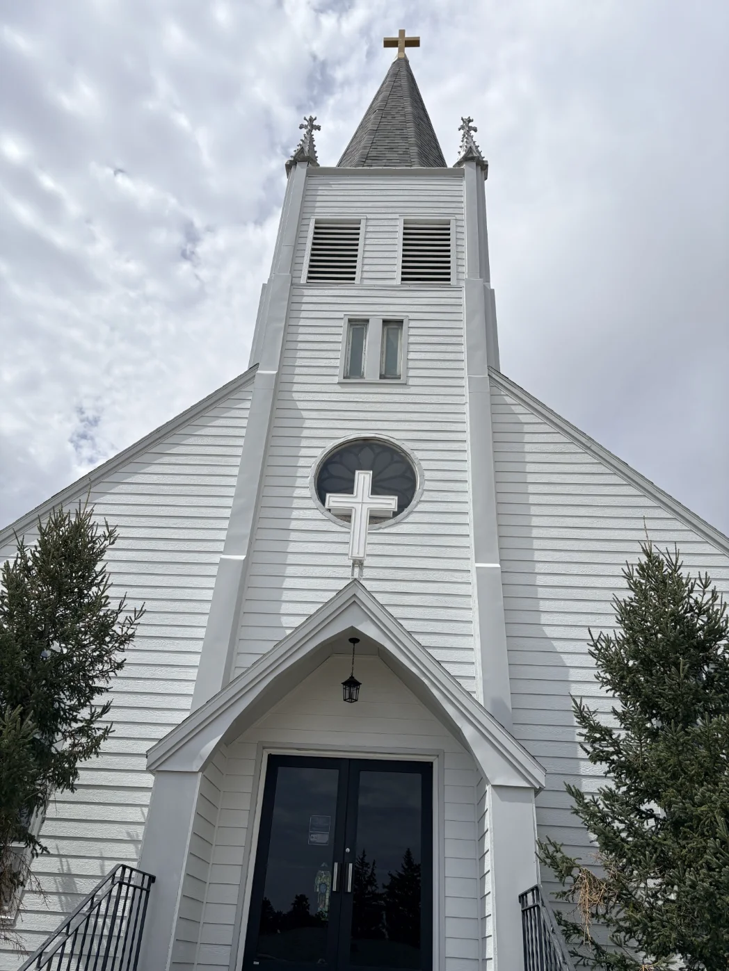 White church steeple with cross above entrance, viewed looking upward against a cloudy sky.