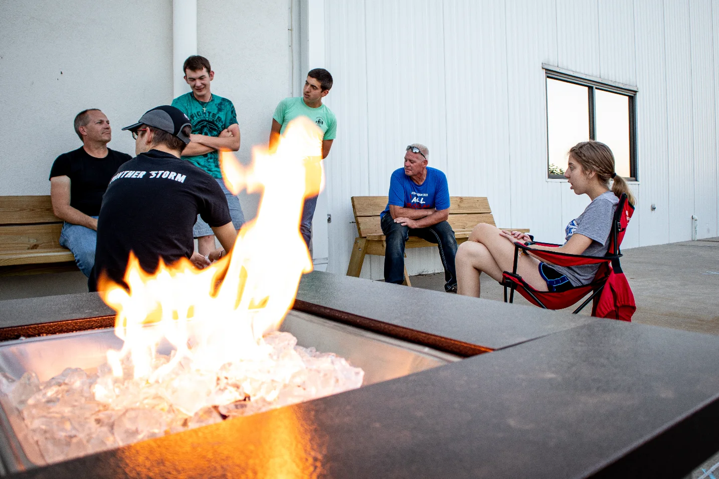 A group of people sitting and standing around a fire pit outside a white building.