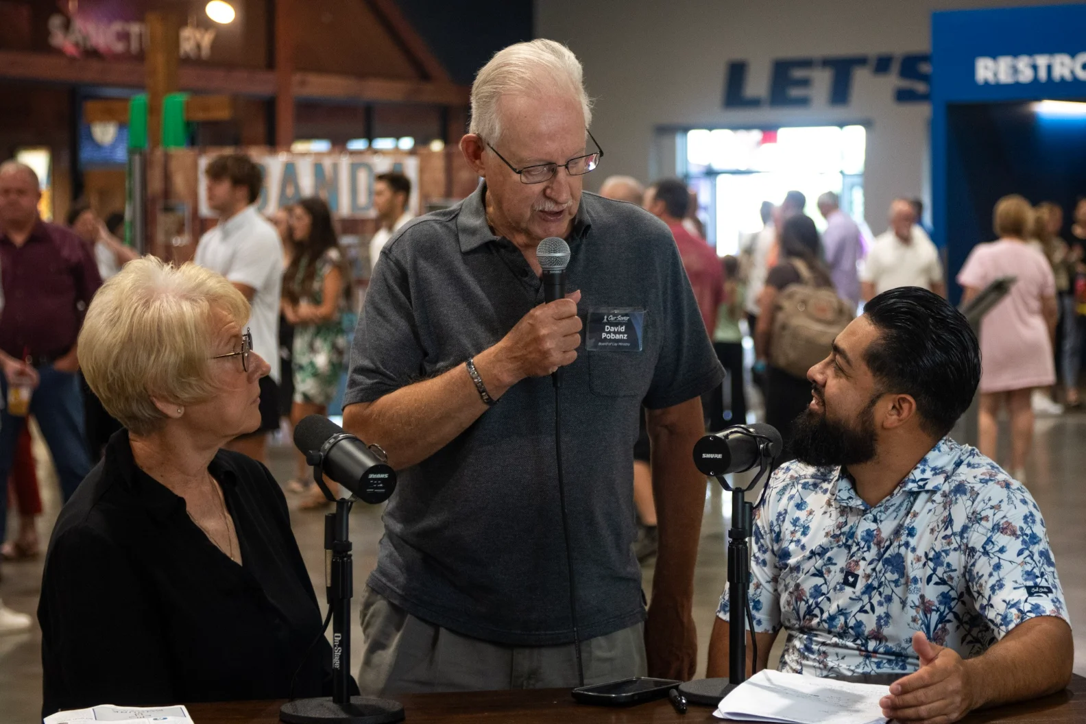Three people engaged in conversation with microphones at an indoor event with others in background.