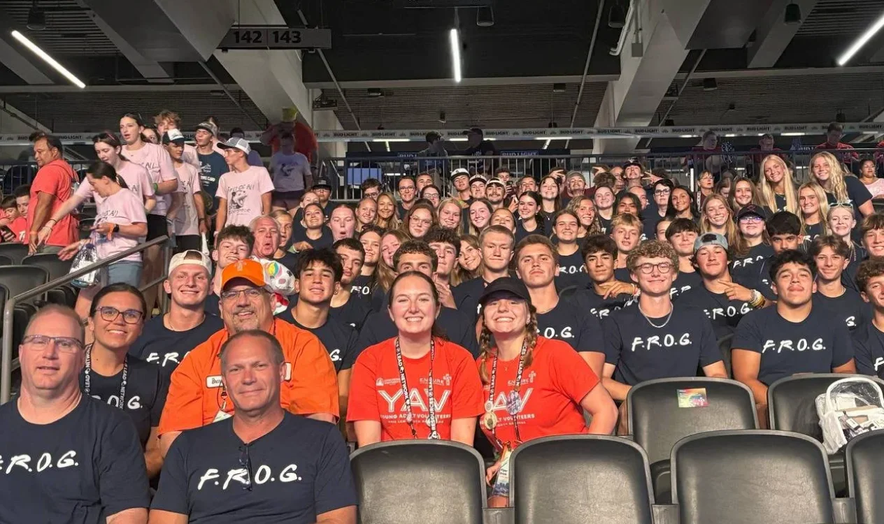 Large youth group seated in stadium seats, many wearing matching F.R.O.G. shirts and two in orange YAV shirts.