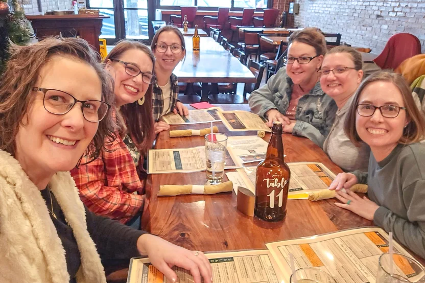 Six women smiling at a restaurant table with menus, glasses, and a Table 11 bottle in the center.