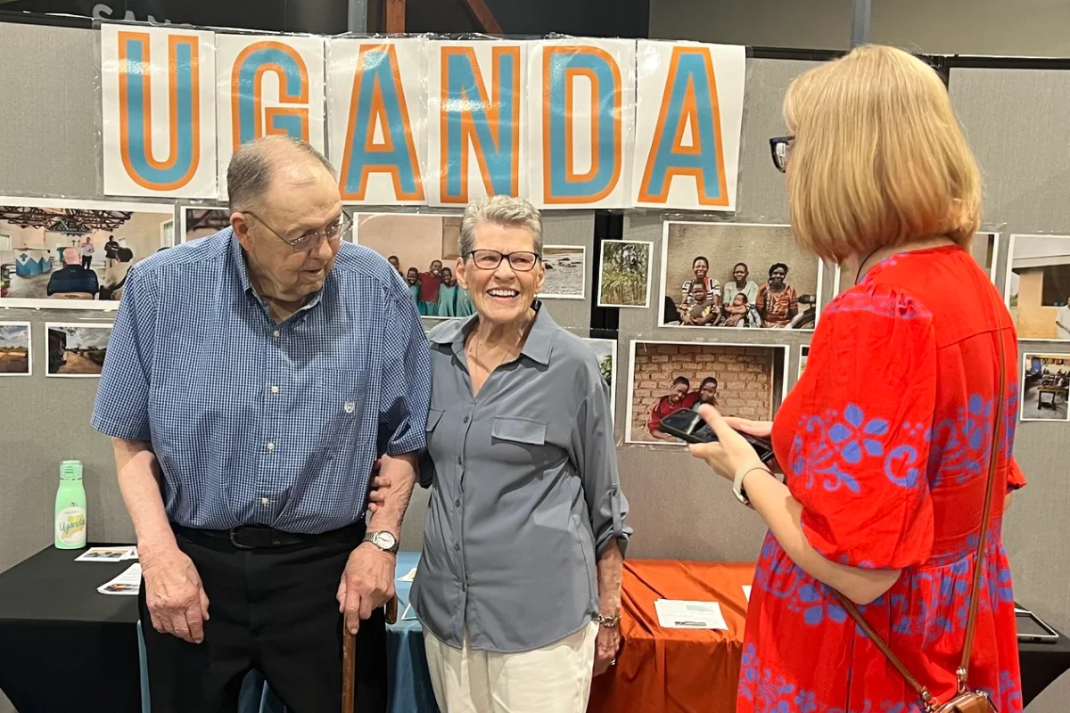 Two elderly people smiling by a Uganda display booth while a woman in a red dress looks at her phone.
