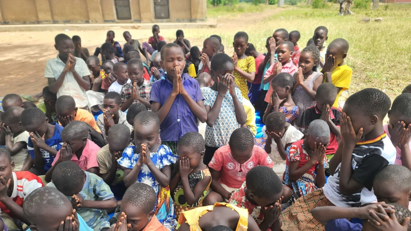 Large group of African children sitting outdoors in prayer with hands clasped or covered faces.