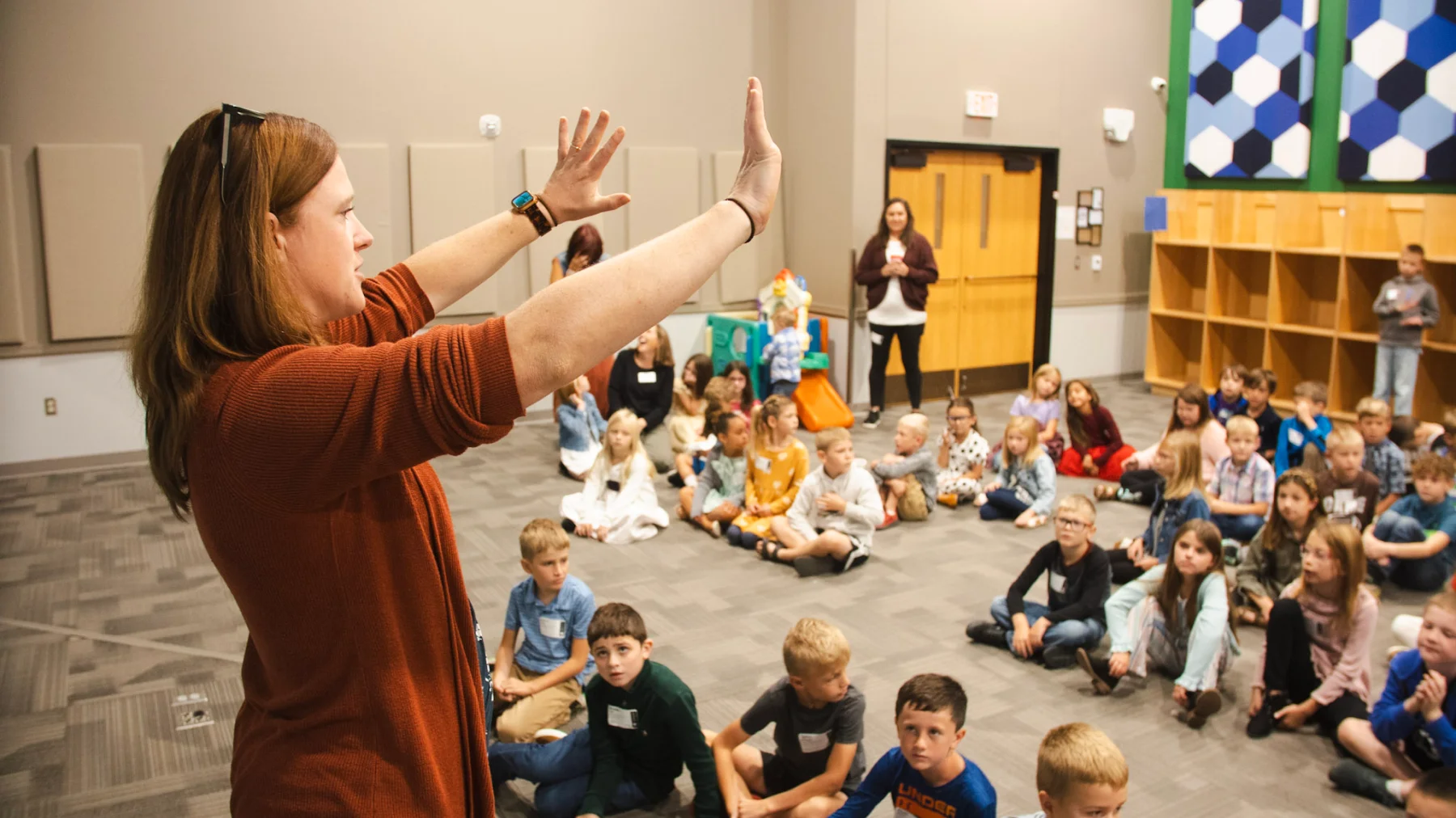Teacher with raised hands leading large group of children sitting on floor in a classroom.