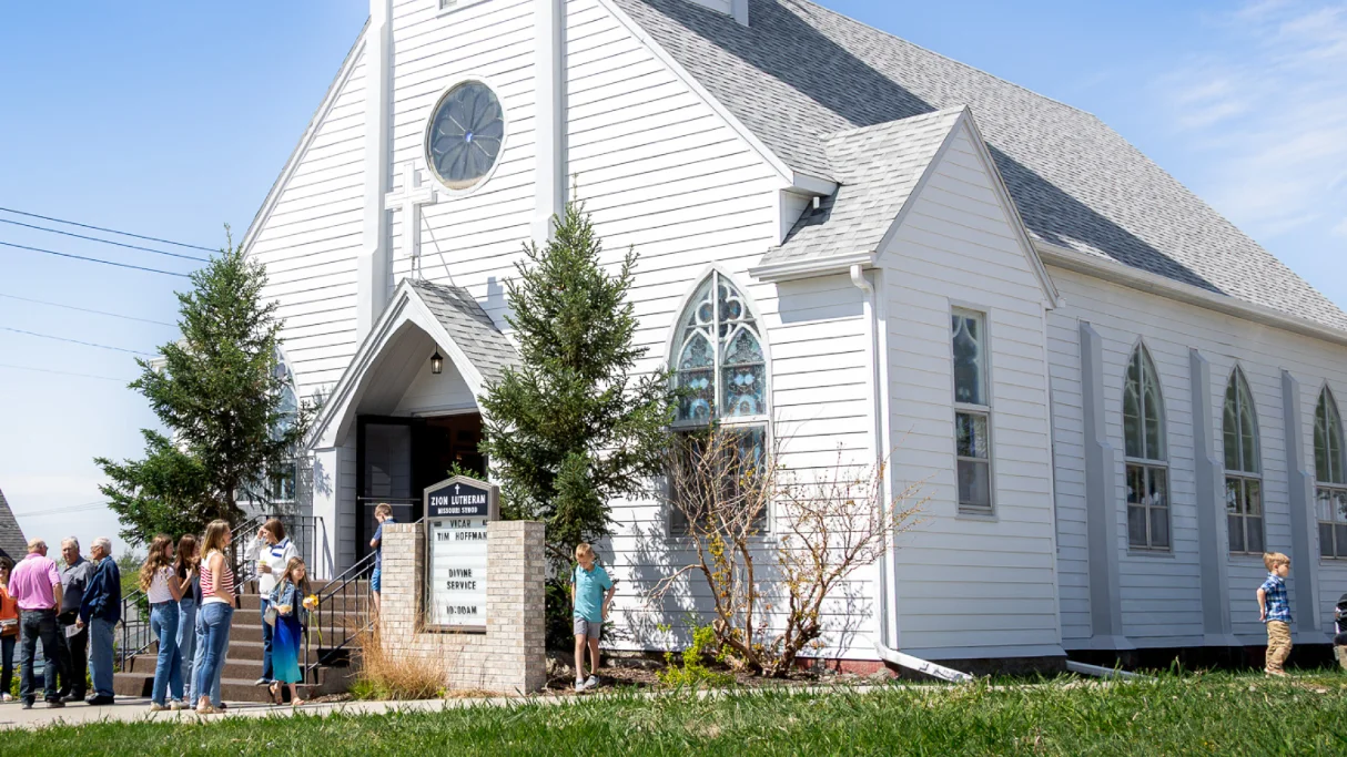 White historic church with stained glass, cross, and people gathered outside on a sunny day.