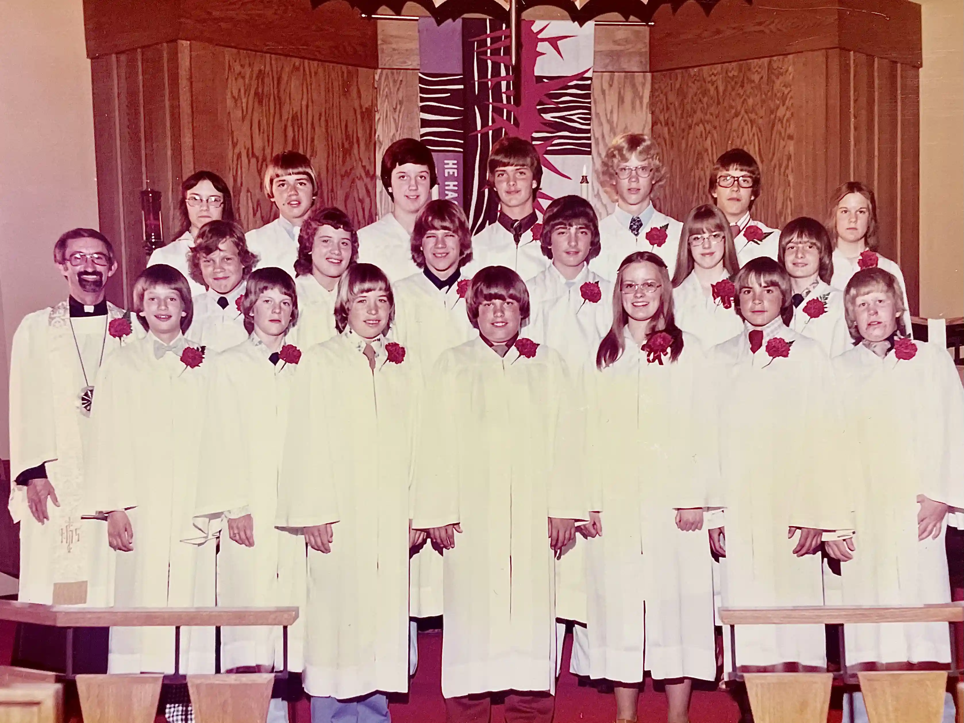 Confirmation class in white robes with red carnations stands with their pastor at the altar.