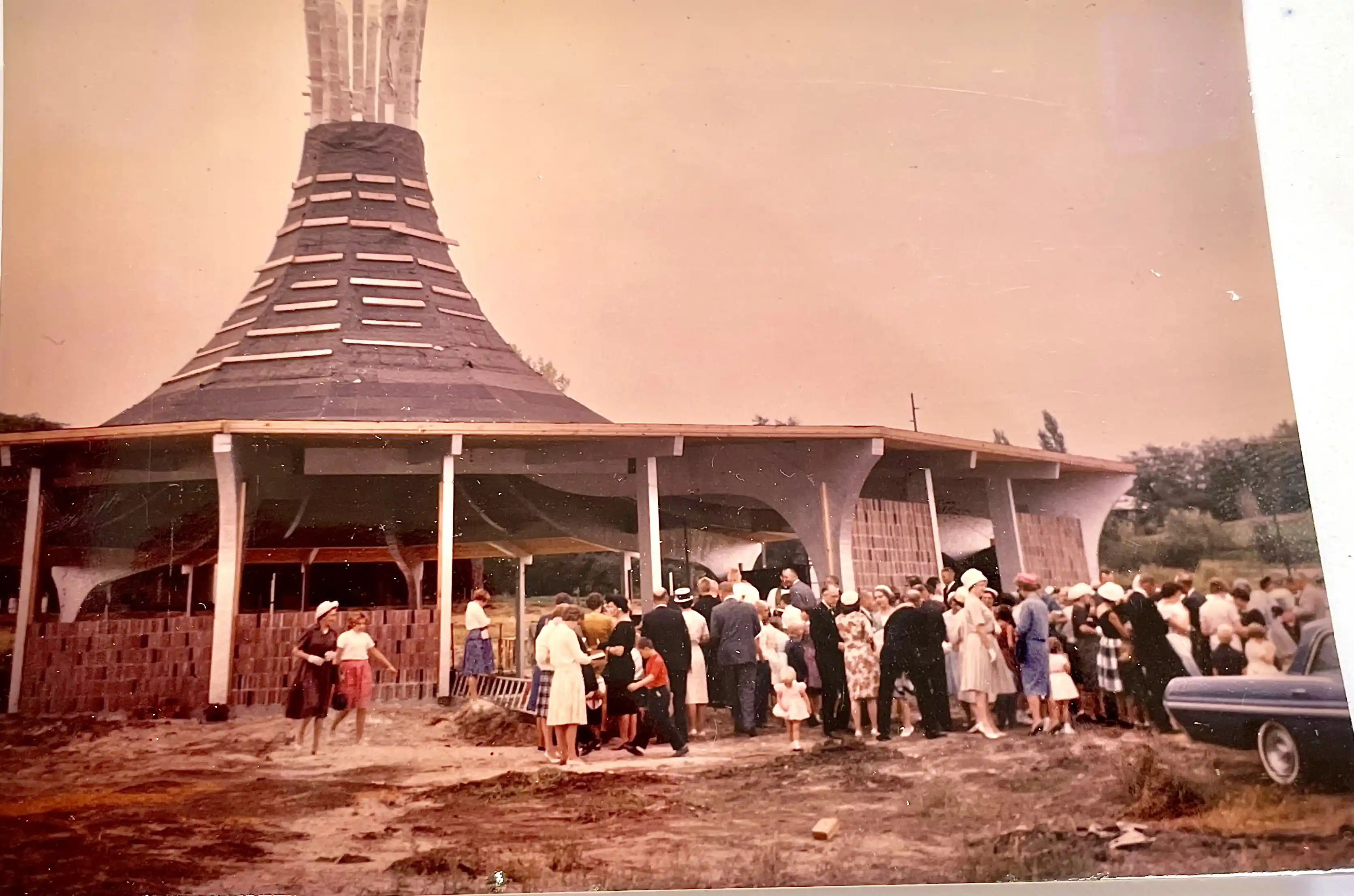 Crowd gathers at the original circular church building with its conical roof under construction.