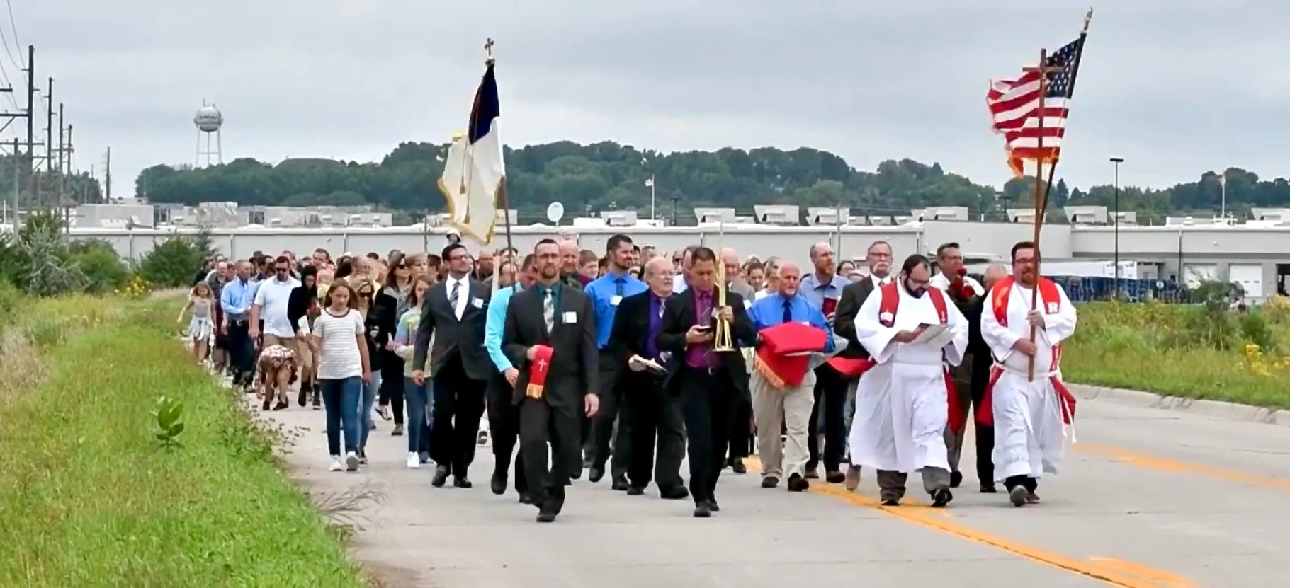 Congregation processes down a road behind pastors carrying Christian and American flags.