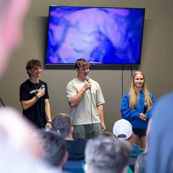 Three youth share testimony with microphones in front of a screen as peers listen.