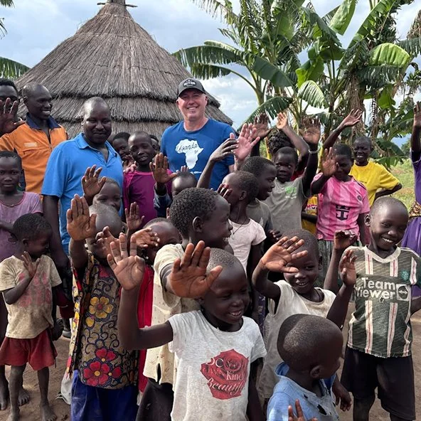Man and village children wave and smile by a thatched hut during the Uganda mission.