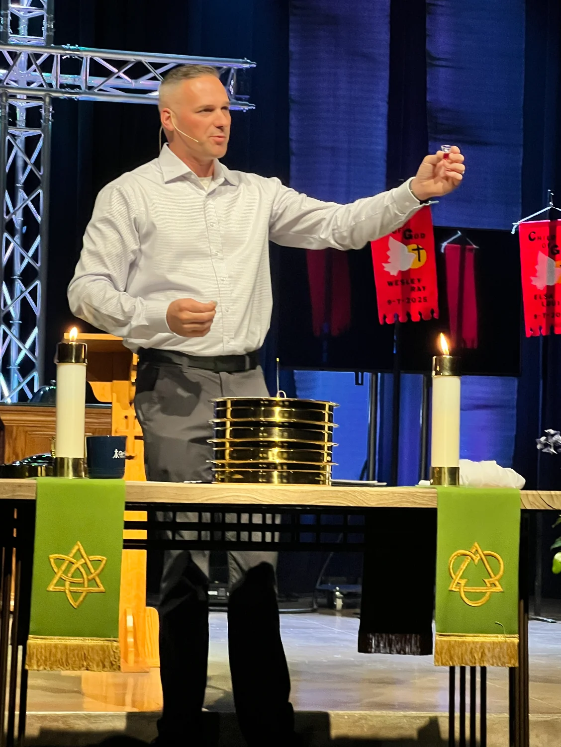 Pastor lifts a communion cup behind an altar with lit candles and stacked communion trays.