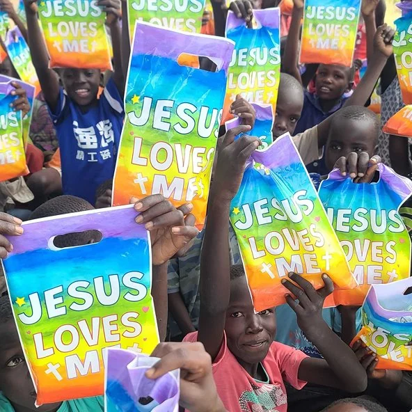 Children hold up colorful Jesus Loves Me gift bags during the Uganda mission outreach.