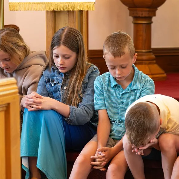 Children sit in a pew with heads bowed and hands folded in prayer.