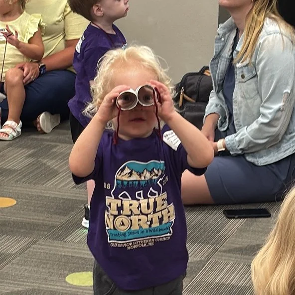 Toddler in a True North VBS shirt peers through handmade craft binoculars.