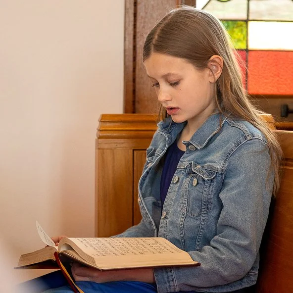 Girl sings from a hymnal in a wooden pew beside a stained glass window.