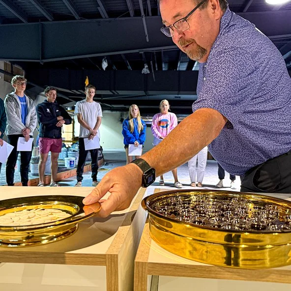 Pastor prepares communion bread and cups on gold trays as youth look on.