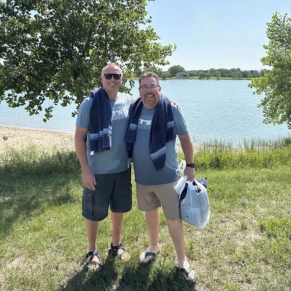 Two men with towels smile by a lakeside beach on a sunny day outing.