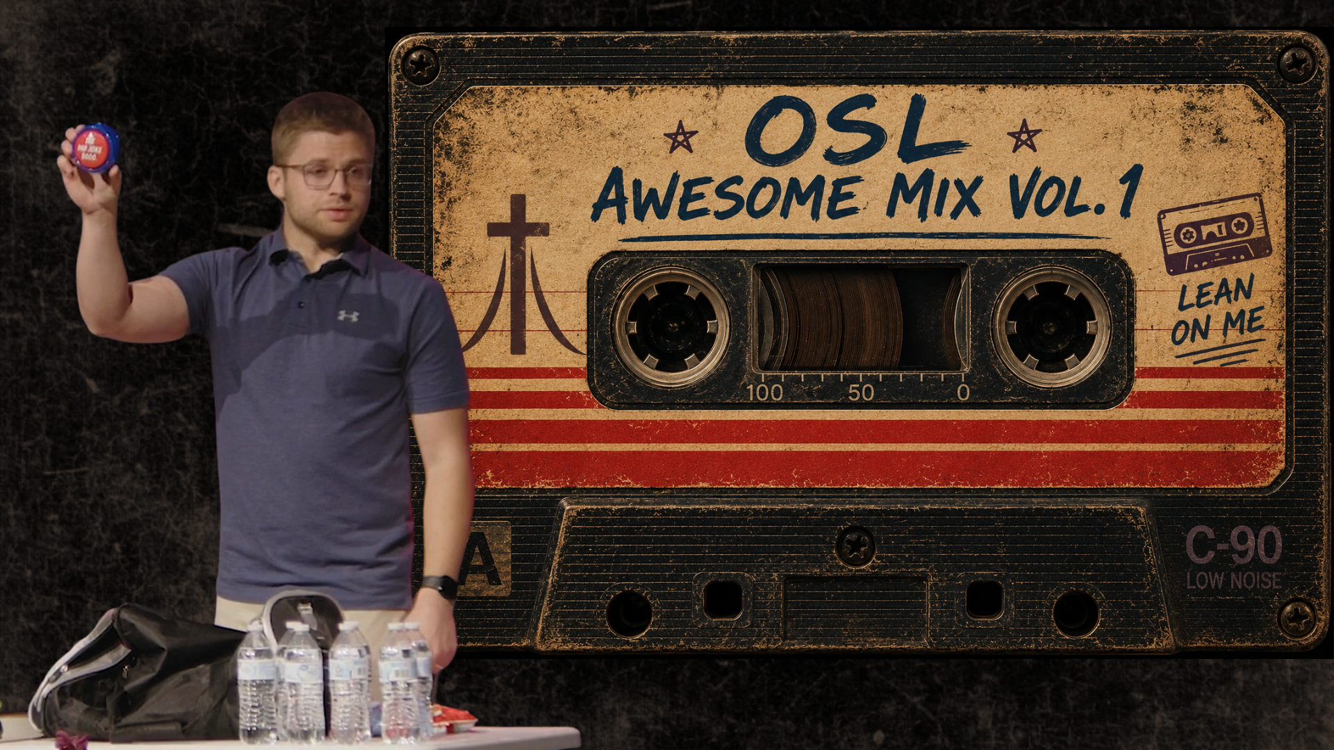 Pastor Caleb holding up a Dad Joke button in front of a backdrop with features a cassette tape with the label OSL Awesome Mix Vol. 1 Lean on Me