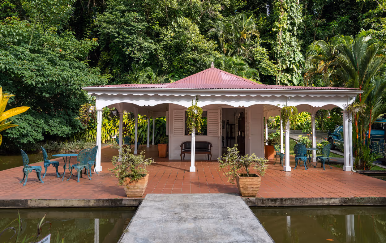Maison de jardin blanche avec toit rouge entourée de végétation luxuriante, avec mobilier extérieur en métal bleu sur une terrasse au bord de l'eau.