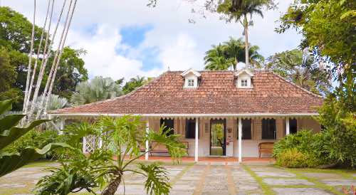 Maison créole avec toit en tuiles et jardin tropical verdoyant sous un ciel bleu partiellement nuageux.
