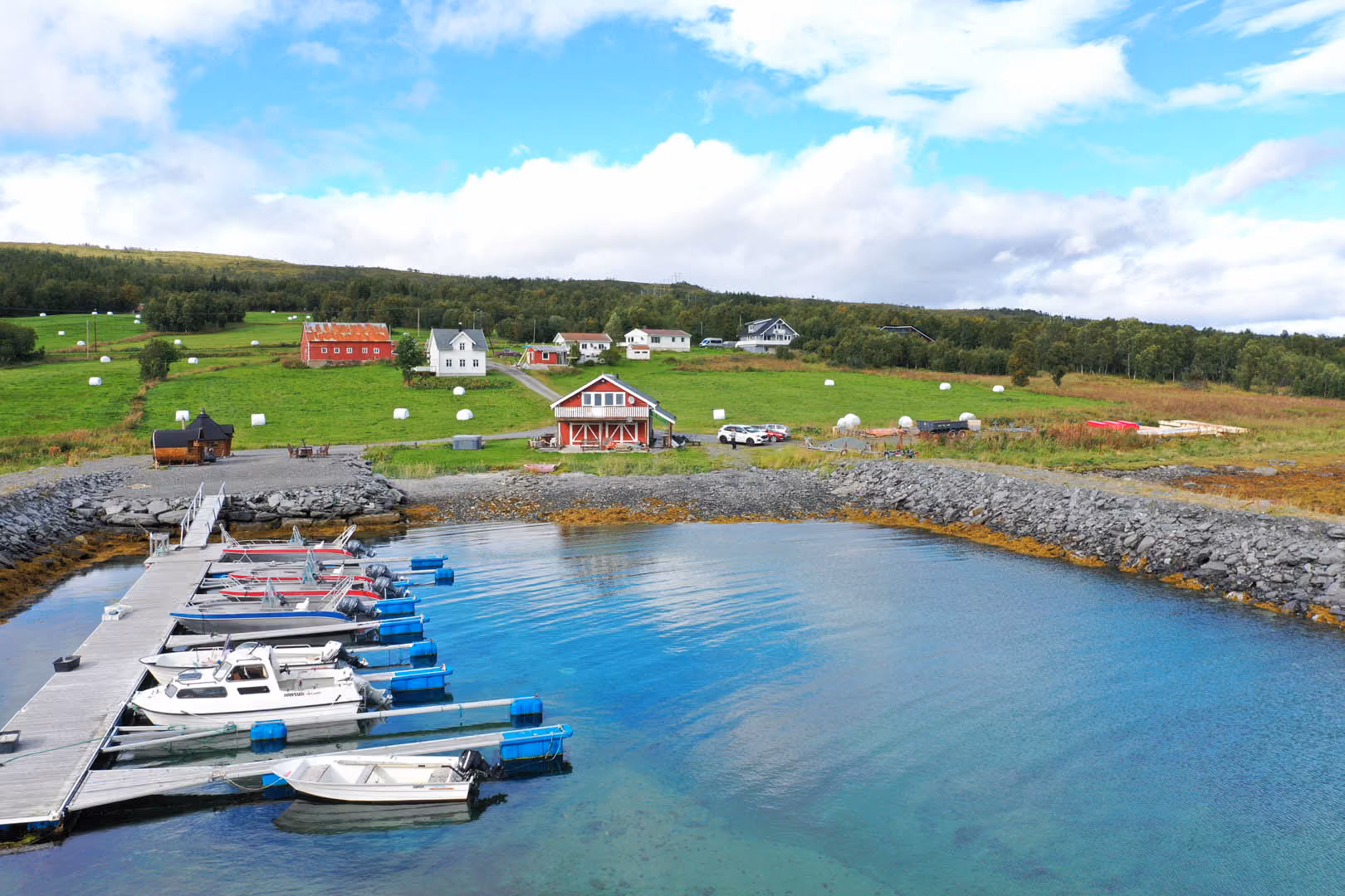 Small boats docked at wooden pier with houses, green fields, and hills under a partly cloudy sky.