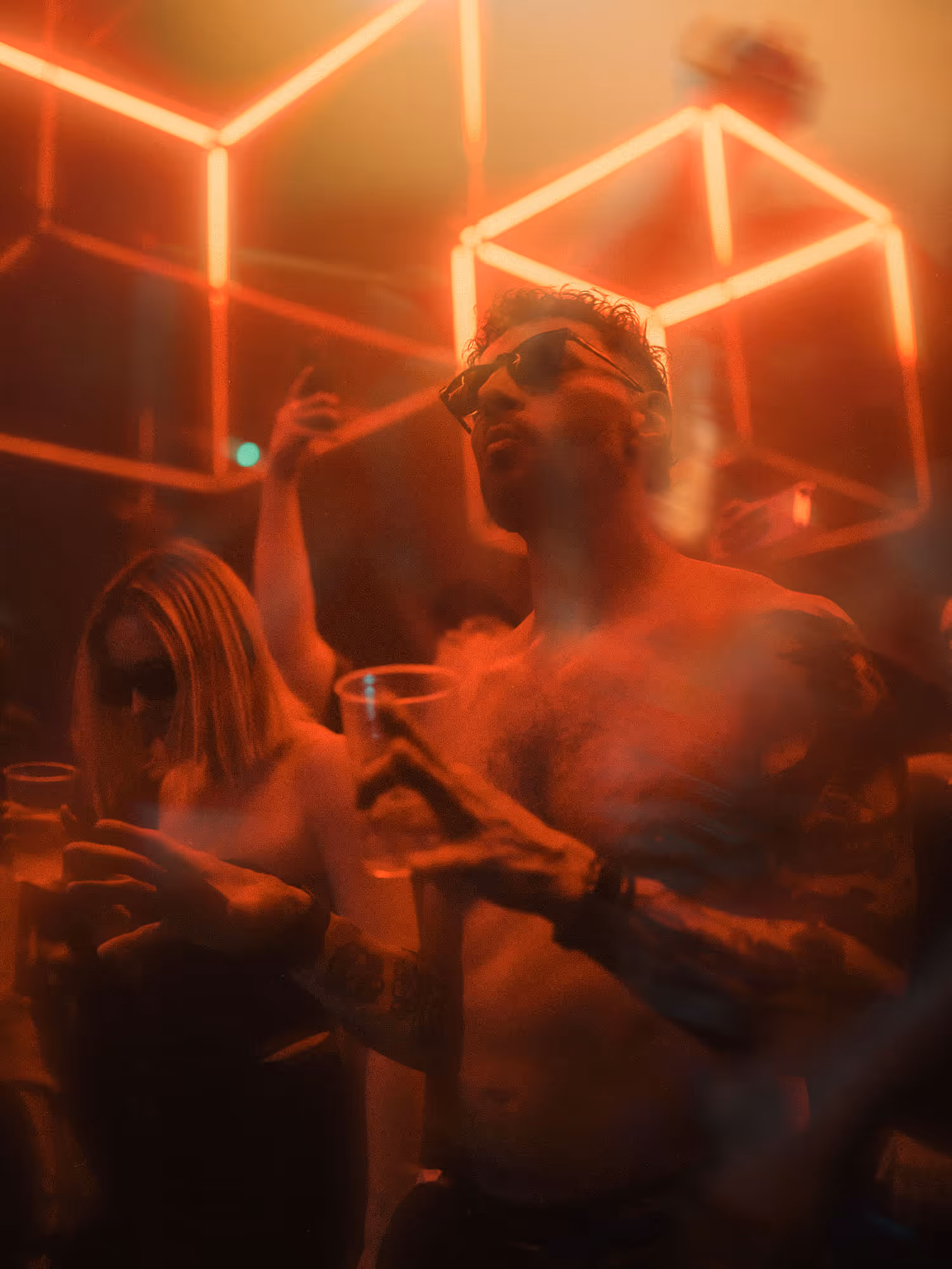 Shirtless man with tattoos and sunglasses holding a drink at a red-lit club with geometric lights.
