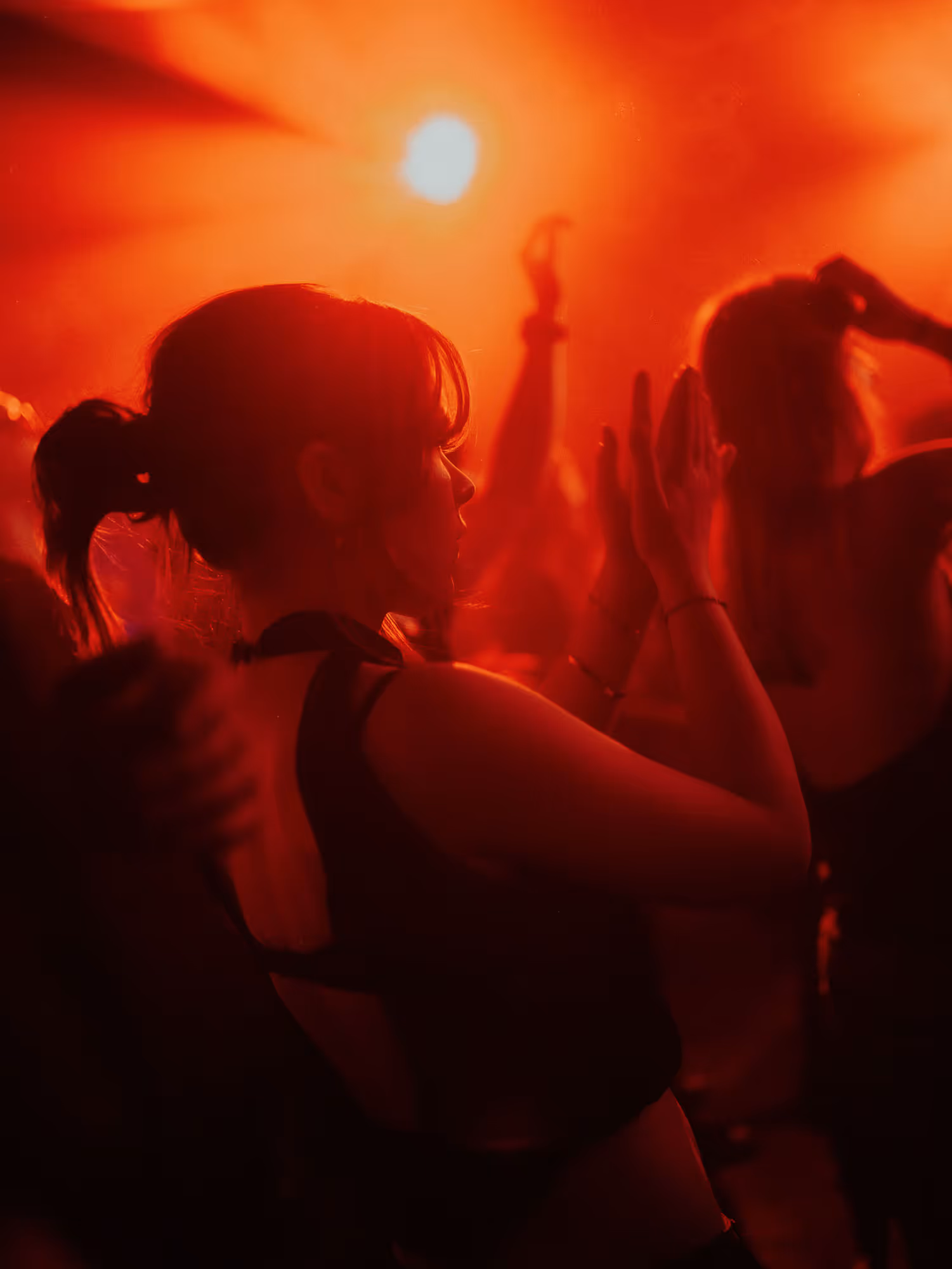 Woman with ponytail clapping hands at a red-lit concert or nightclub with crowd around her.