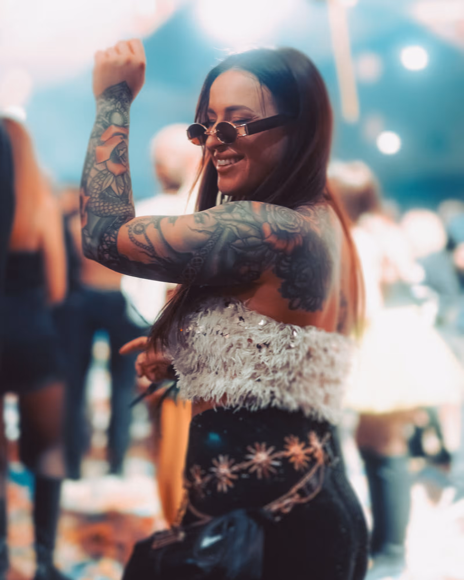Smiling woman with tattoos dancing, wearing sunglasses and a white fuzzy top at a lively event.