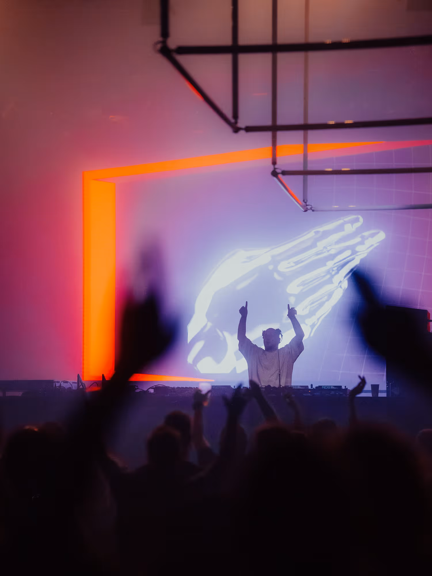 DJ performing with raised hands in front of crowd and glowing skeletal hand visuals on screen.