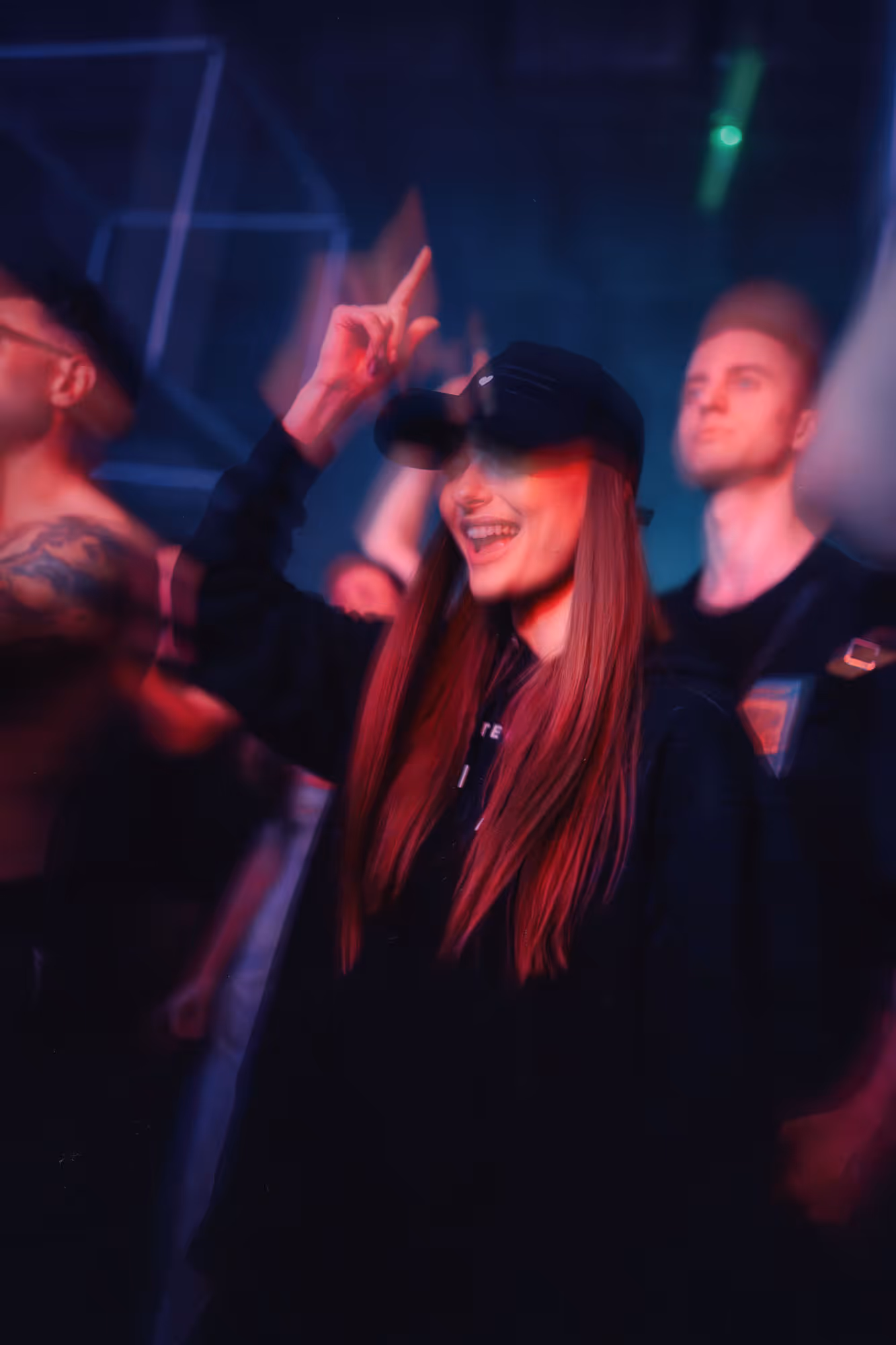 Smiling woman in black cap and hoodie dancing with raised hand at a dimly lit concert or club.