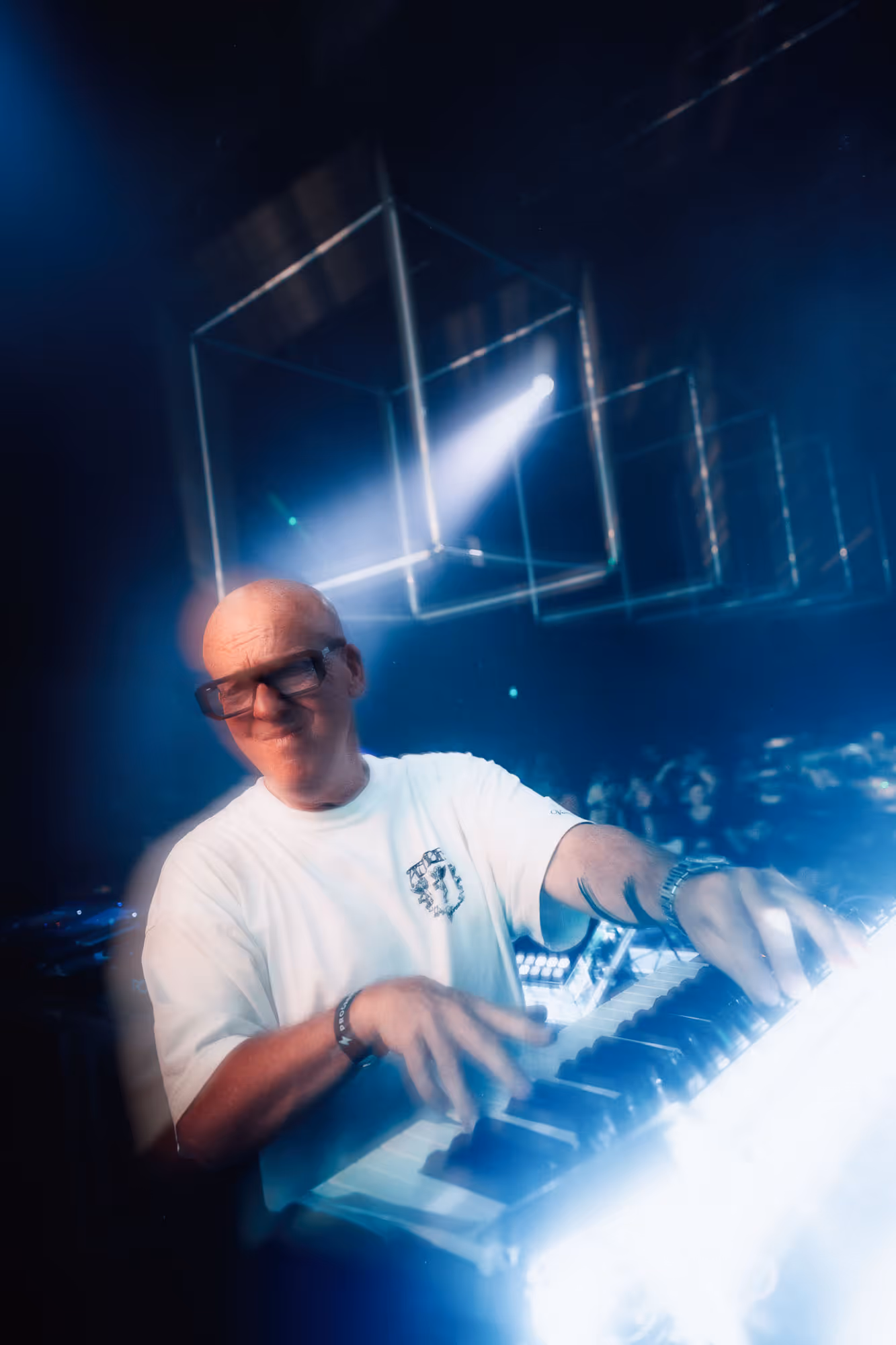 Man wearing glasses playing keyboard on stage with light cubes hanging and blue lighting effects.