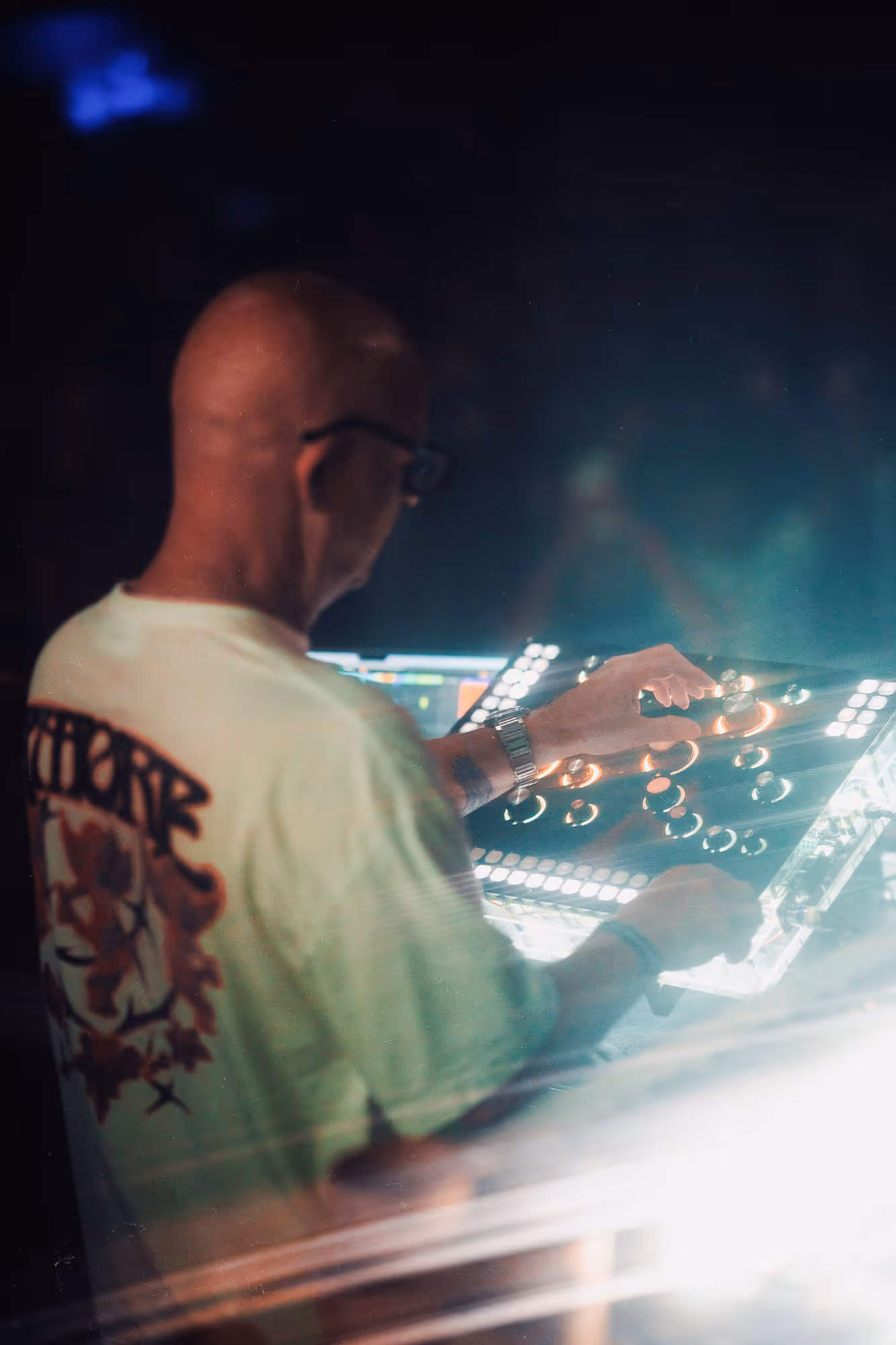 Bald man wearing glasses adjusts knobs on illuminated DJ mixer during a live event at night.