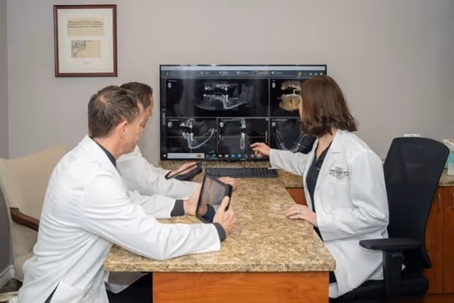 Three dentists in white coats reviewing dental x-rays displayed on a monitor in an office.