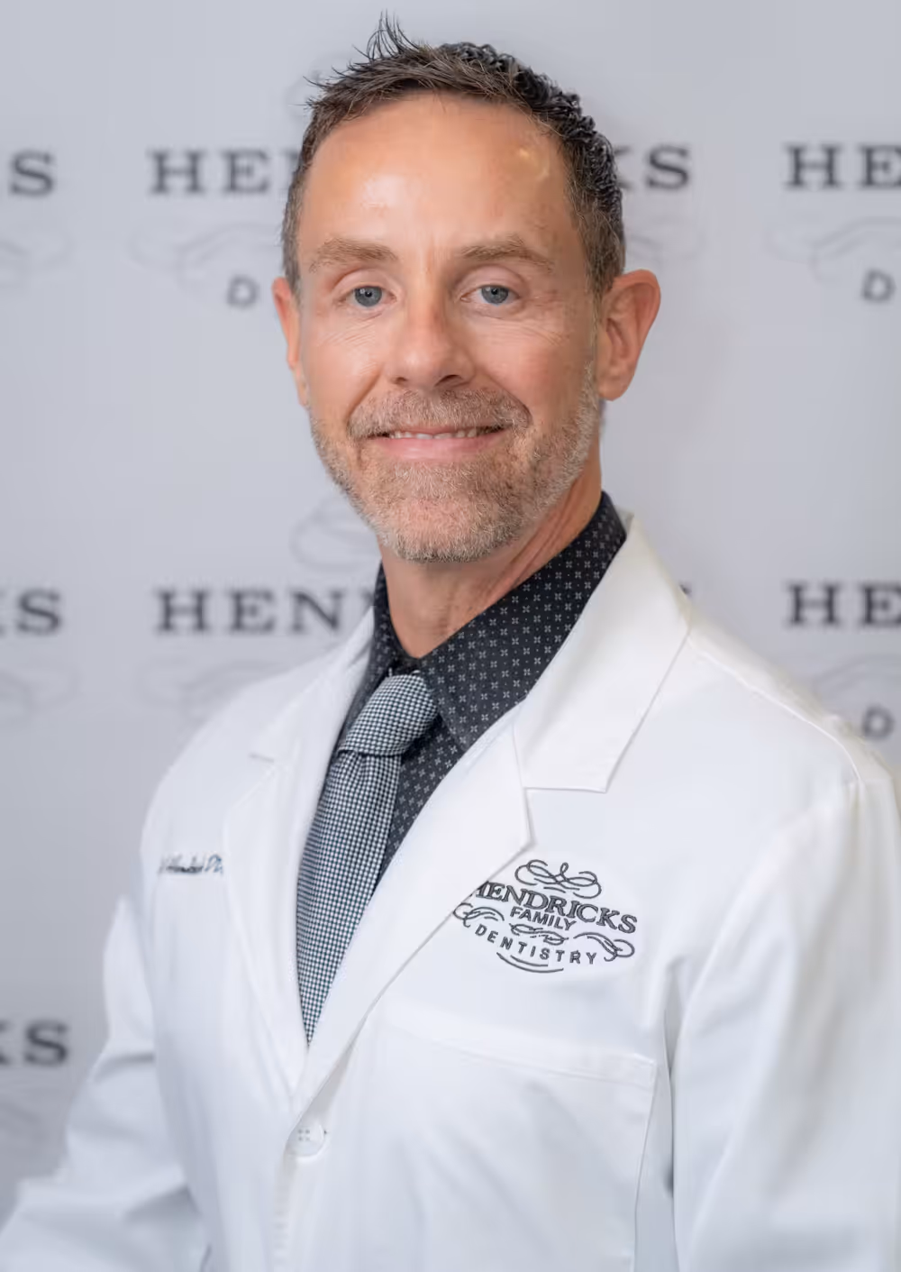 Smiling male dentist in a white coat with Hendricks Family Dentistry logo on it.
