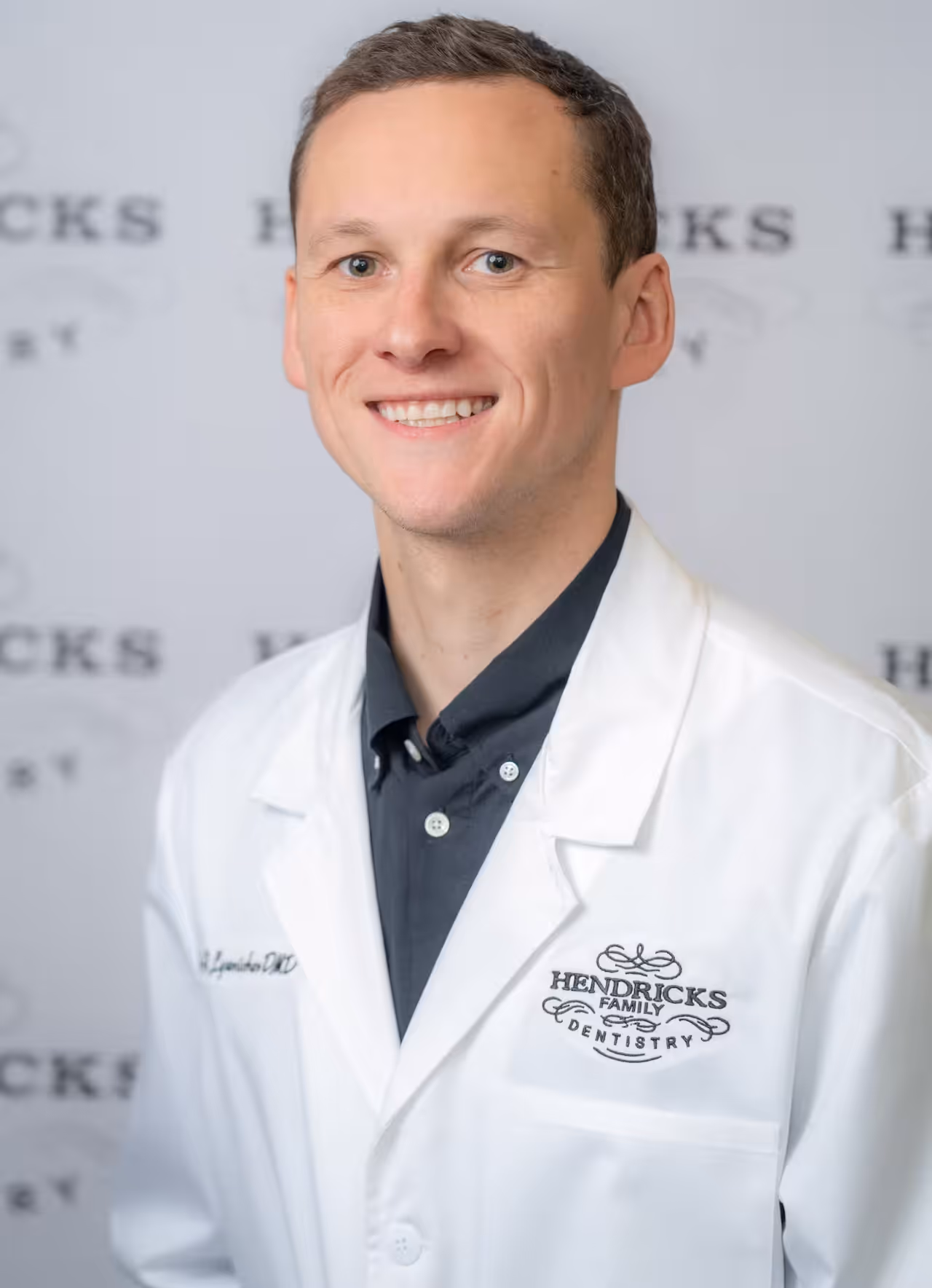Smiling man in white lab coat with Hendricks Family Dentistry logo on chest.