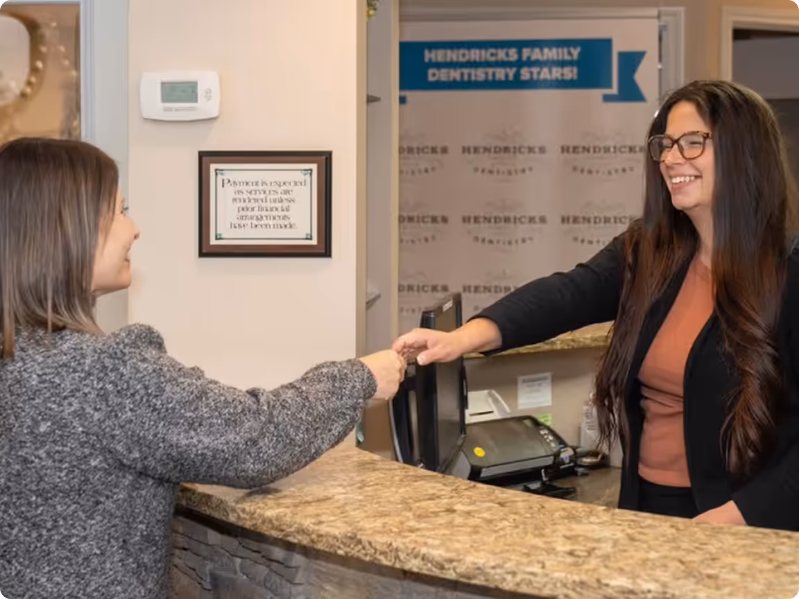 Receptionist with glasses smiling and handing a keycard to a woman at a dental office counter.