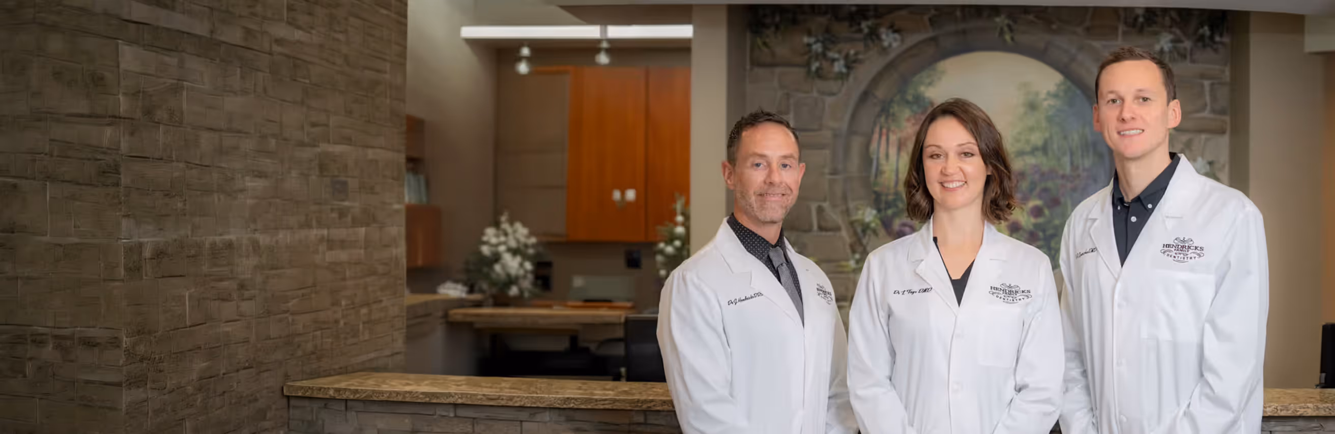 Three dentists in white coats smiling inside a modern dental office reception area.