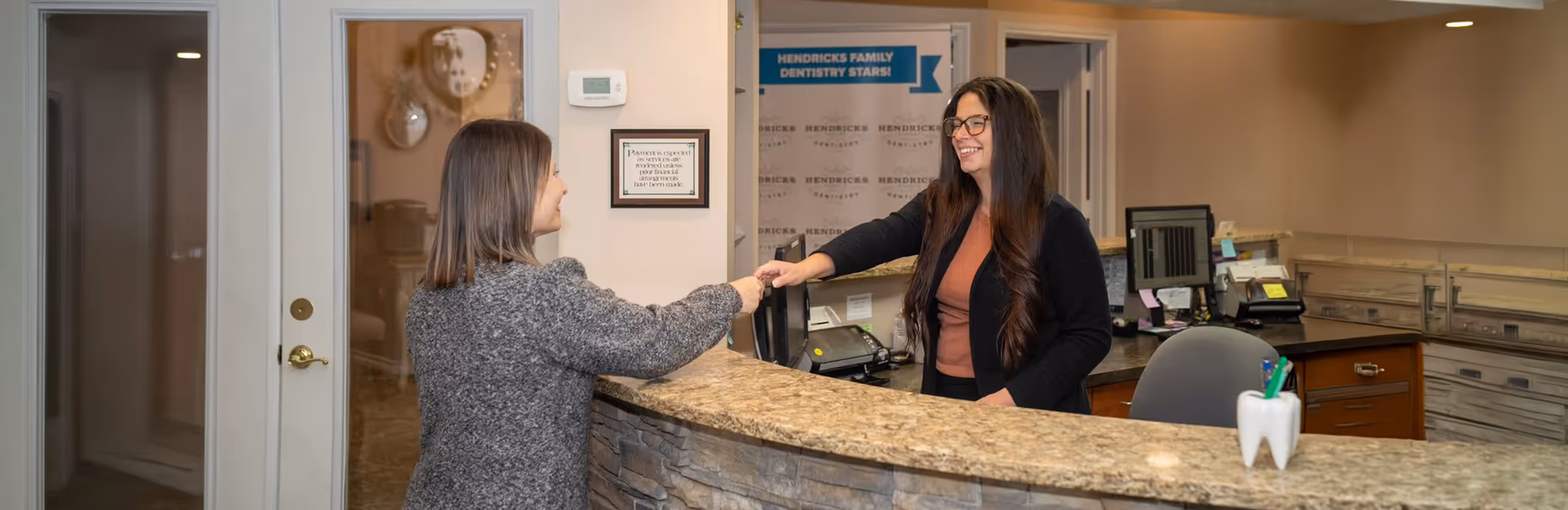 Receptionist smiling and handing a key card to a woman at a dental office front desk.