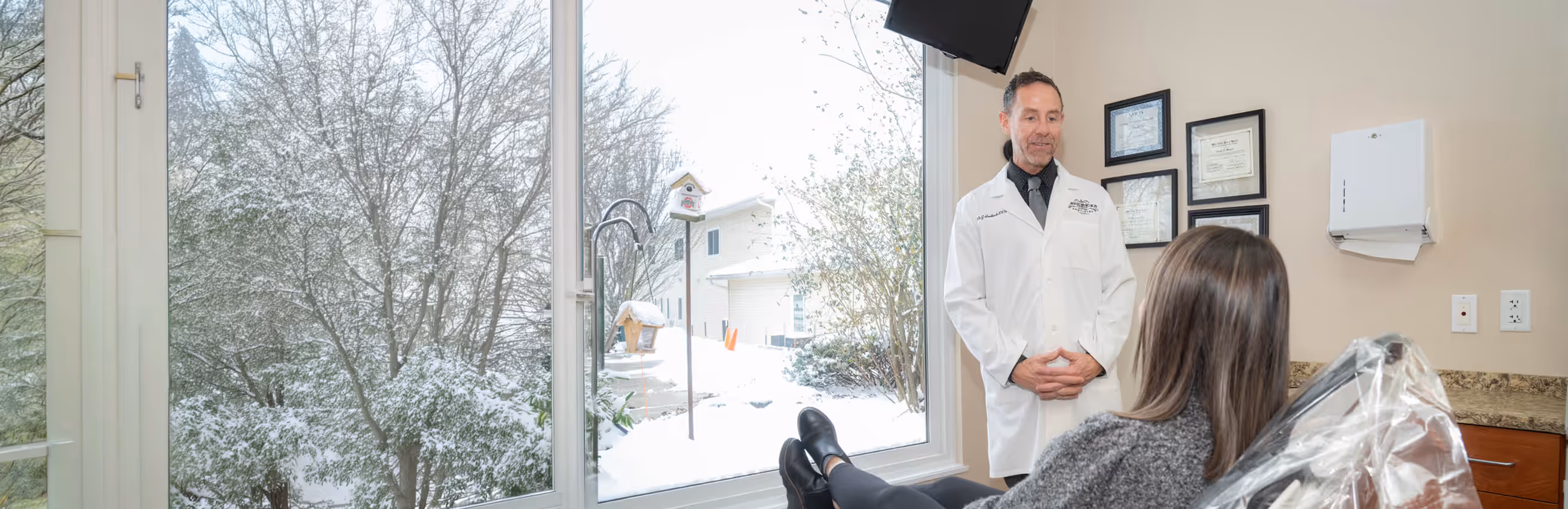 Dentist in white coat talking to female patient seated in dental chair by a large window with snow outside.