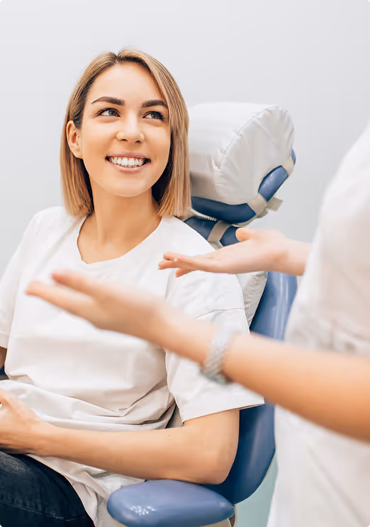 Smiling woman sitting in a medical chair listening to another person speaking with hand gestures.
