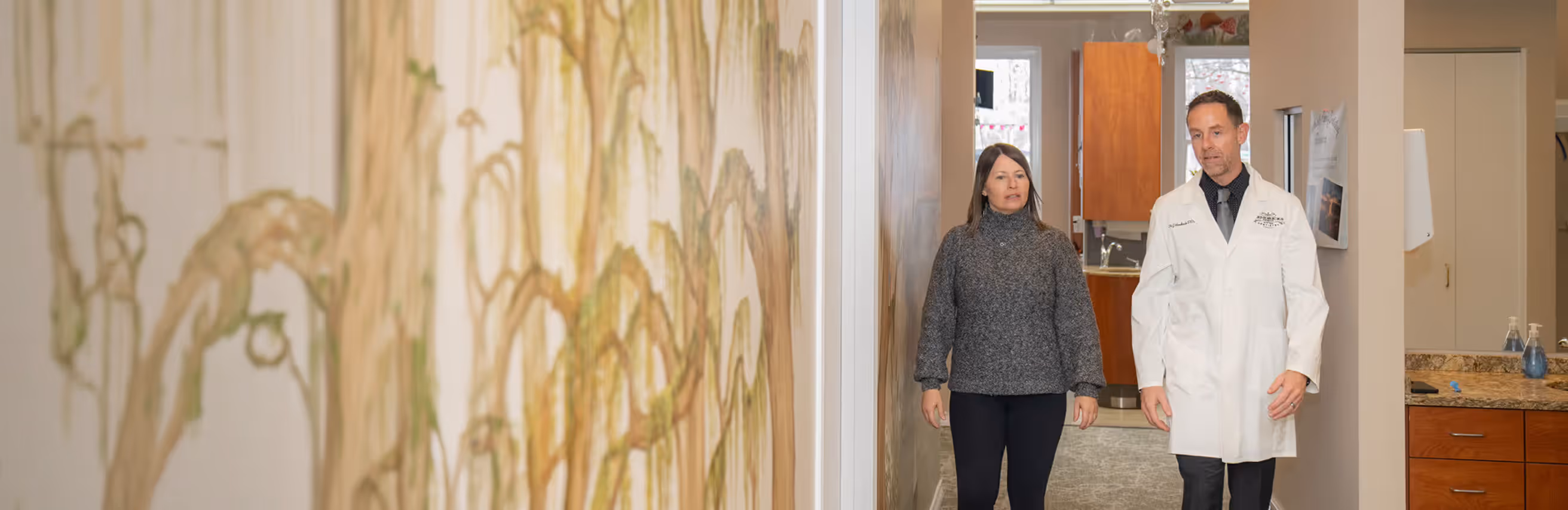 Doctor in white coat walking and talking with woman in gray sweater in a hallway.